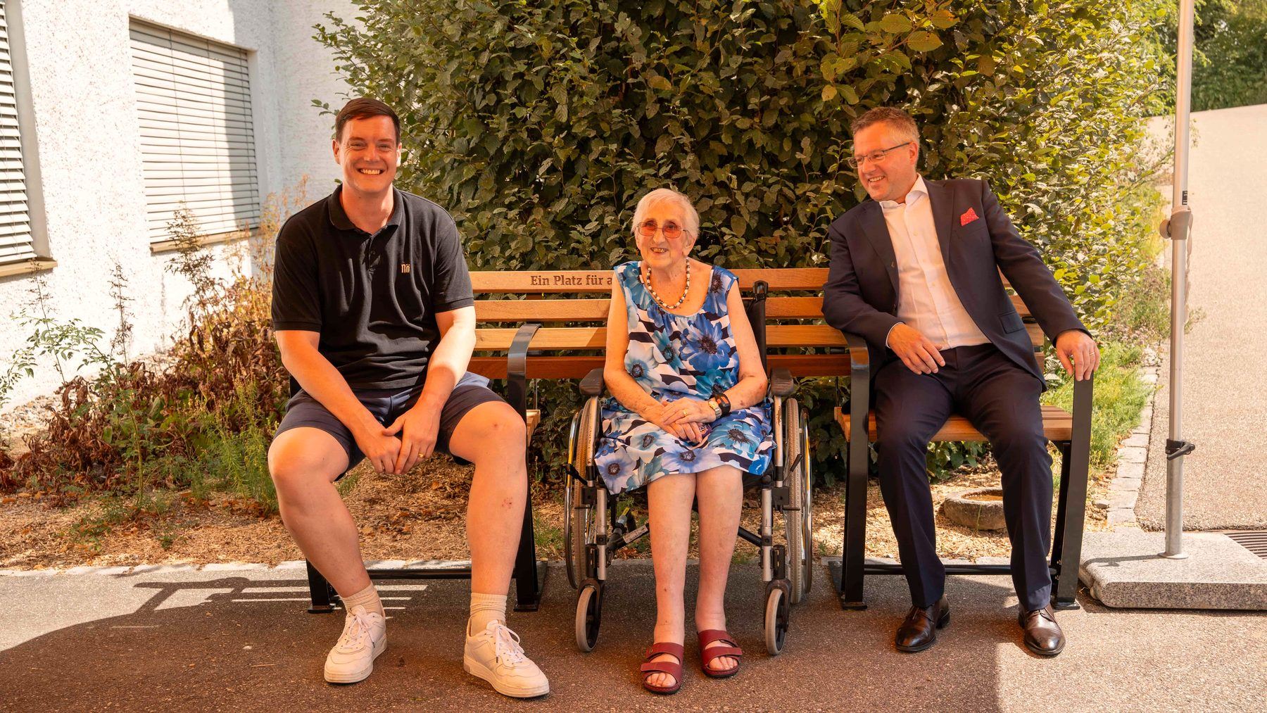 Drei Menschen sitzen nebeneinander auf einem Inklusionsbänkli – eine Person im Rollstuhl, integriert in der Mitte